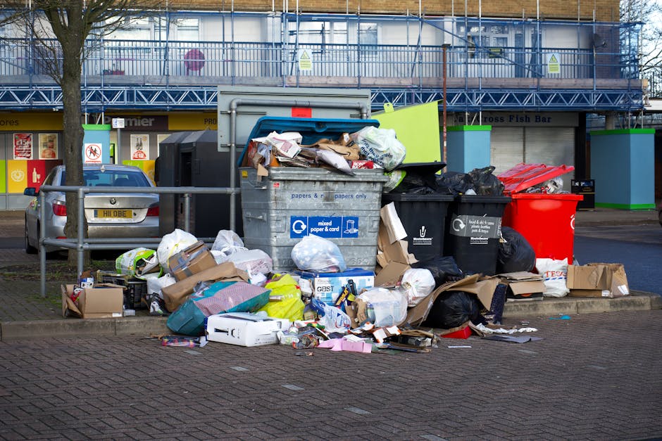 A patch of ground near a fence line is littered with discarded cardboard boxes, some flattened and torn, along with crumpled paper, plastic packaging, and other miscellaneous waste materials scattered