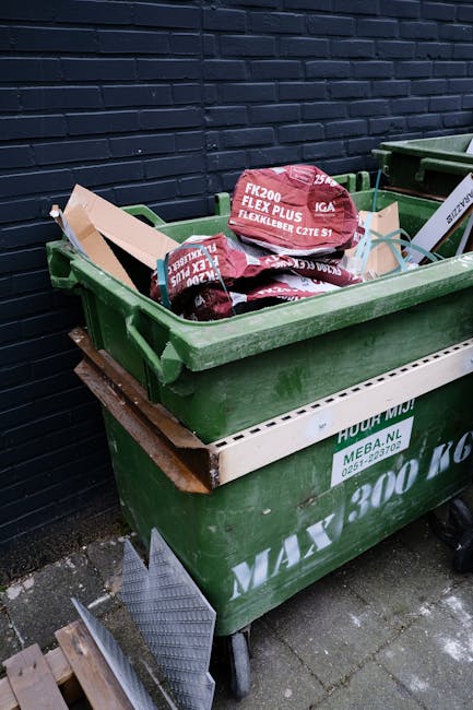 A waste collection worker wearing a blue uniform with an orange reflective vest and a blue cap is emptying a large blue wheeled bin into a white rubbish collection truck parked on a cobblestone street