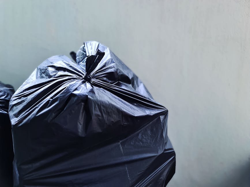 A large green old clothing donation bin located outdoors on a pavement area, surrounded by dense greenery and bushes in the background. The bin has a rectangular opening at the top and four smaller co